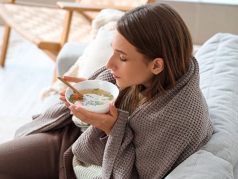 Frau mit Decke um den Schultern, sitzt auf der Couch und isst Suppe.