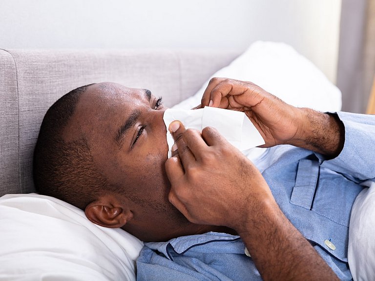 Einer Sinusitis geht häufig ein Erkältungsvirus mit Schnupfen voraus. Mann im Schlafanzug liegt mit einer Sinusitis im Bett.