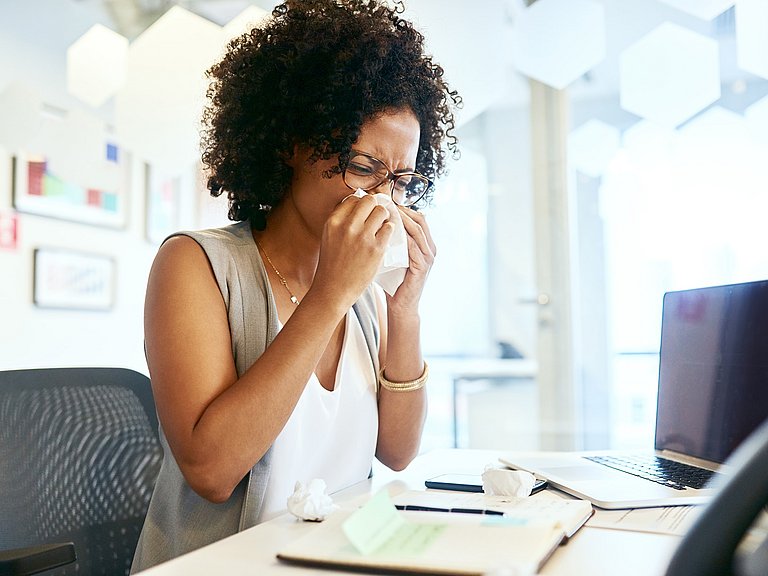 Frau sitzt am Schreibtisch und putzt sich die Nase.