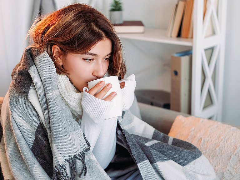 Frau sitzt mit Decke auf der Couch und trinkt Tee.