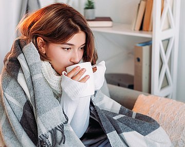 Frau sitzt mit Decke auf der Couch und trinkt Tee.