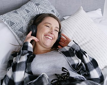 Frau mit Erkältung und Schnupfen liegt im Bett und hört einen Podcast.