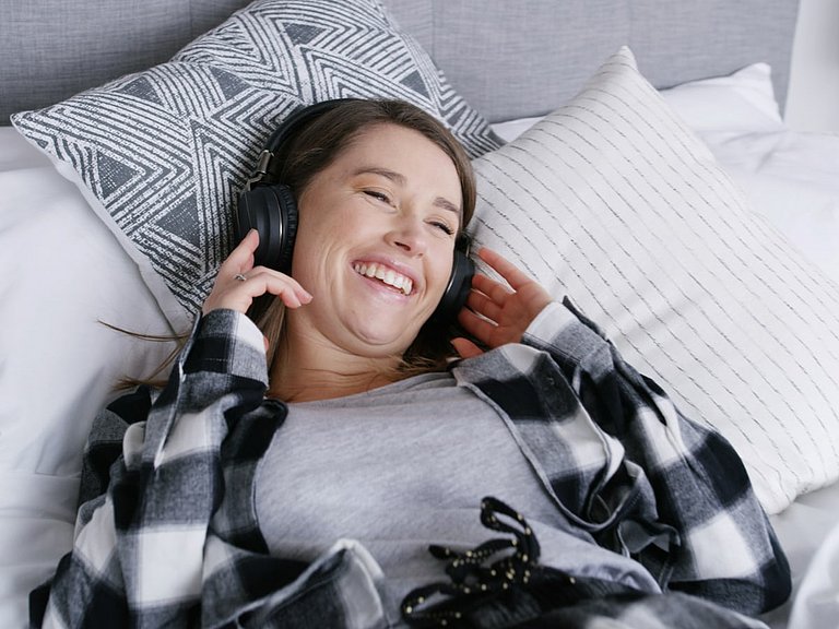 Frau mit Erkältung und Schnupfen liegt im Bett und hört einen Podcast.