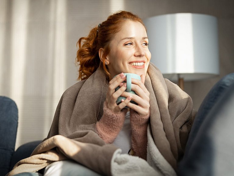 Frau mit Wolldecke und Teetasse auf einem Sofa. 