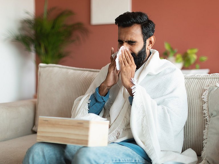 Wenn die Nase ständig läuft, wird das auch als Fließschnupfen bezeichnet. Mann auf der Couch mit Fließschnupfen putzt sich die Nase.