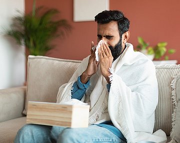 Mann auf der Couch mit Fließschnupfen putzt sich die Nase.