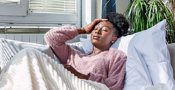 Bei einer Sinusitis können Hausmittel helfen die Symptome zu lindern. Frau liegt mit Decke und Kissen auf einer Coach.