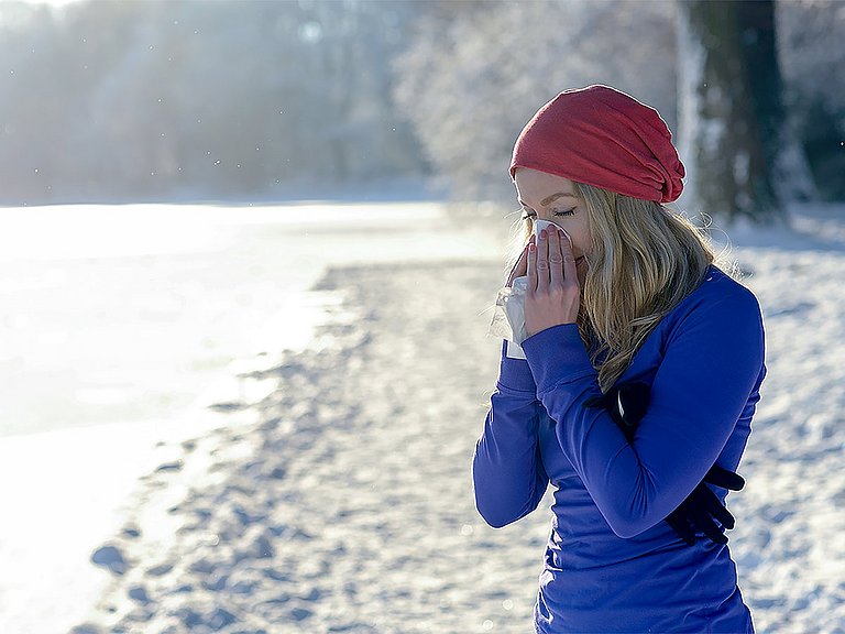Viele möchten trotz Schnupfen nicht auf Sport verzichten. Frau mit Schnupfen macht eine Pause beim Sport im Schnee und putzt sich die Nase.