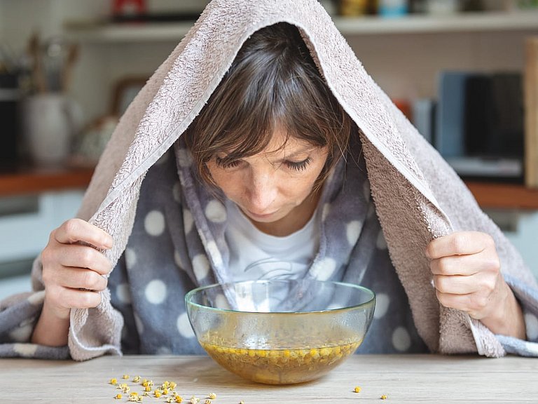 Frau mit Bademantel und Handtuch inhaliert ein Kamillendampfbad.