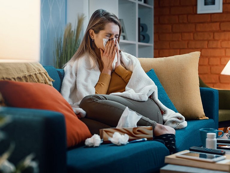 Frau sitzt auf einem Sofa und putzt sich die Nase.